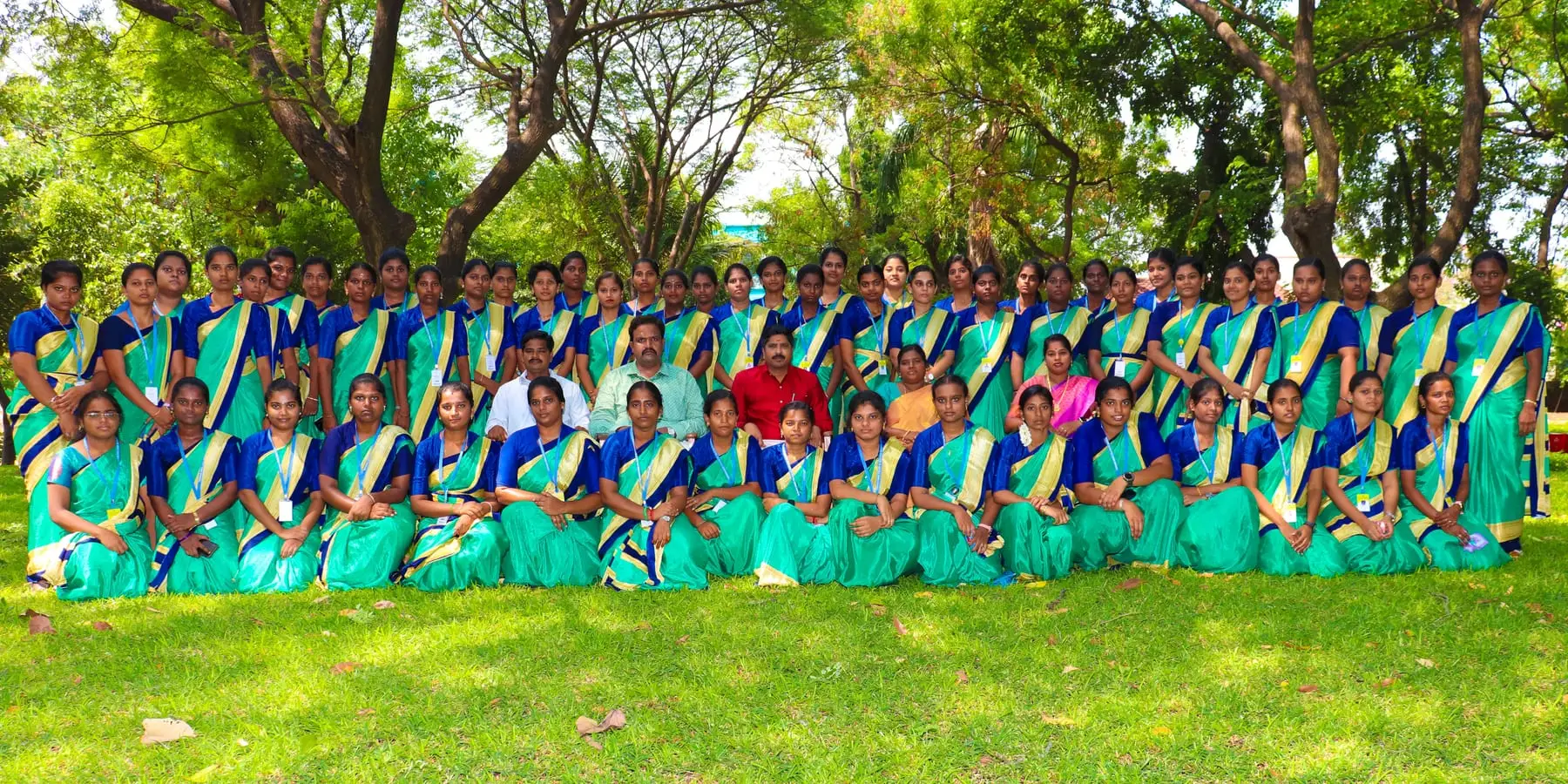 A batch of students in matching green and blue saris poses with faculty on a lawn under trees.