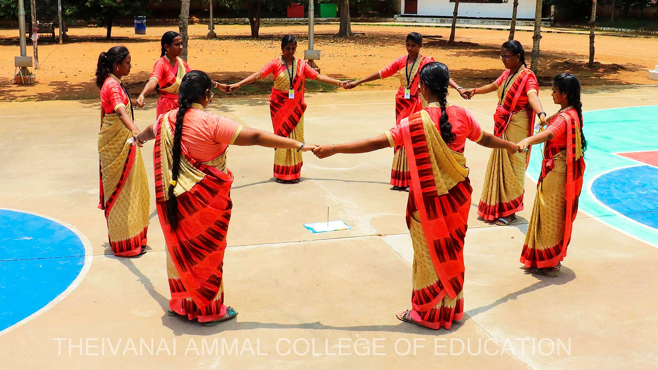 Students in saris hold hands in a circle on an outdoor court.